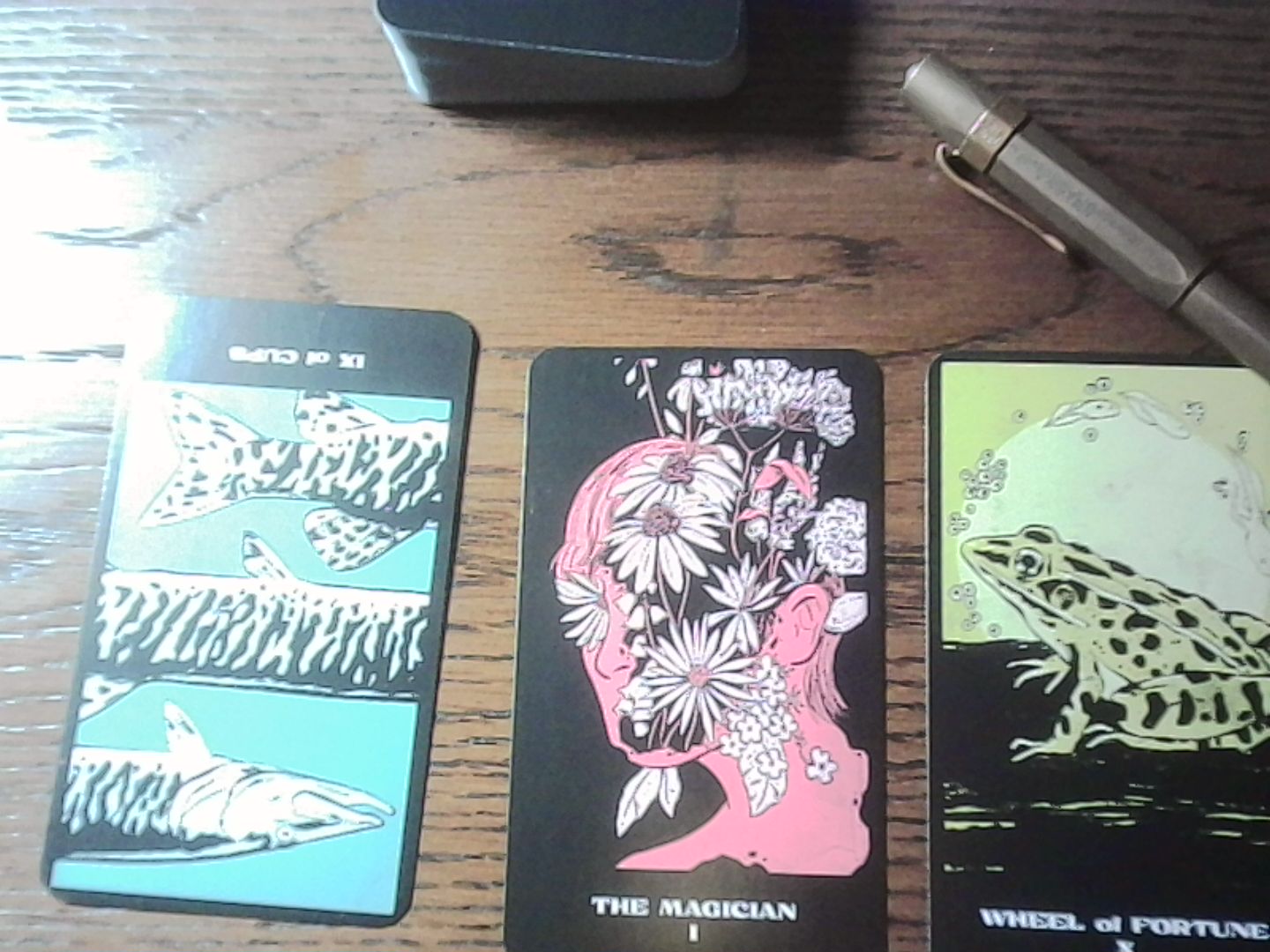 Reversed IX of Cups, The Magicial, and The Wheel Of Fortune spread out on a desk. Cards are from The Great Lakes Tarot.