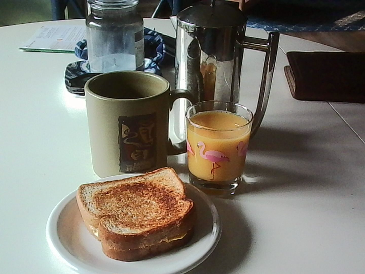 An egg, cheese, and ham sandwich on a plate in front of a coffee mug, glass of orange juice, and french press. All are on a white table.