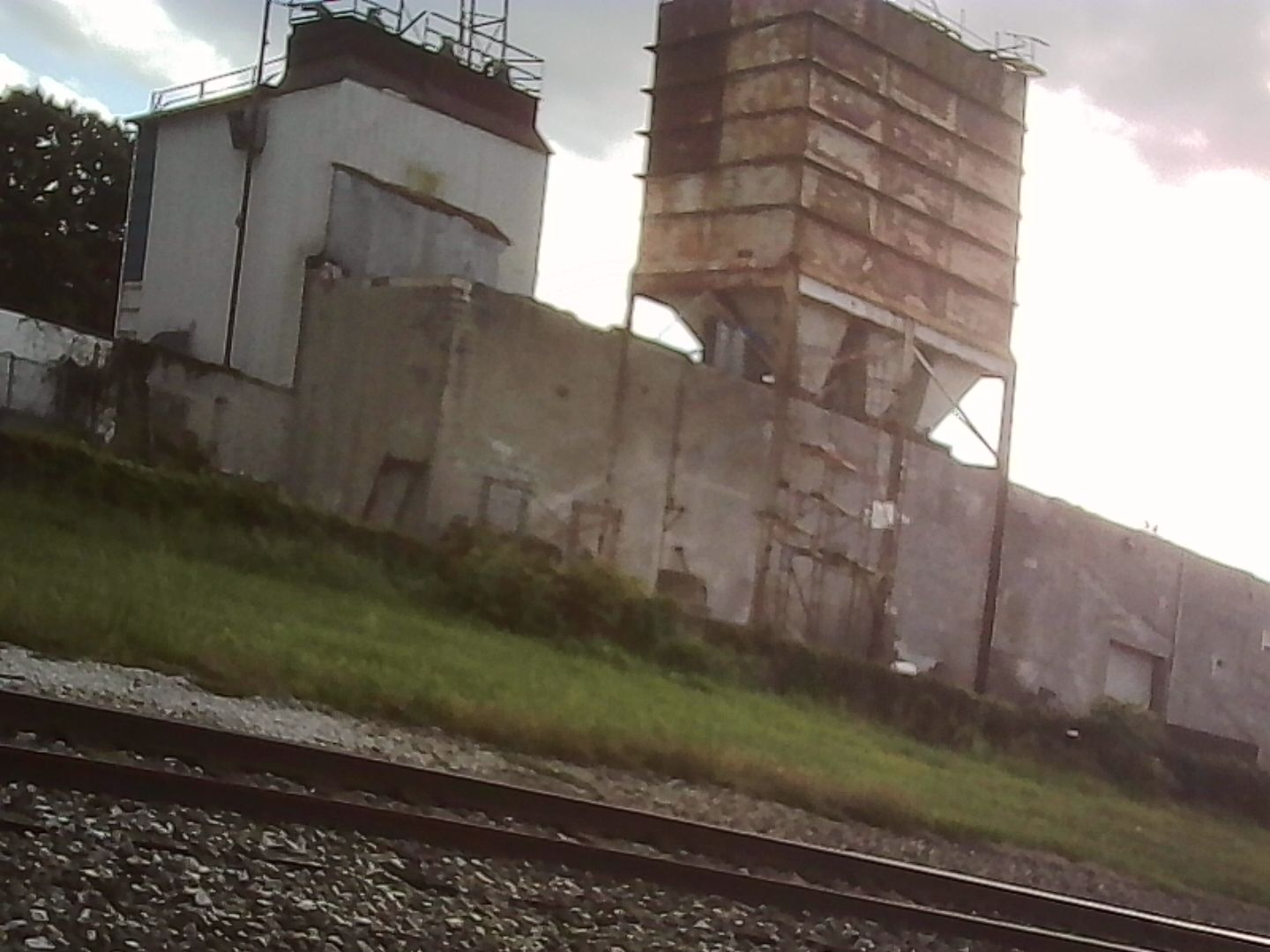 A decaying industrial building next to railroad tracks and a strip of barely contained grass and assorted plant life.